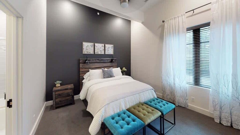 Rest comfortably in this serene bedroom featuring a cozy bed, dark accent wall, soft natural light, and a colorful bench that adds a vibrant touch to the space.