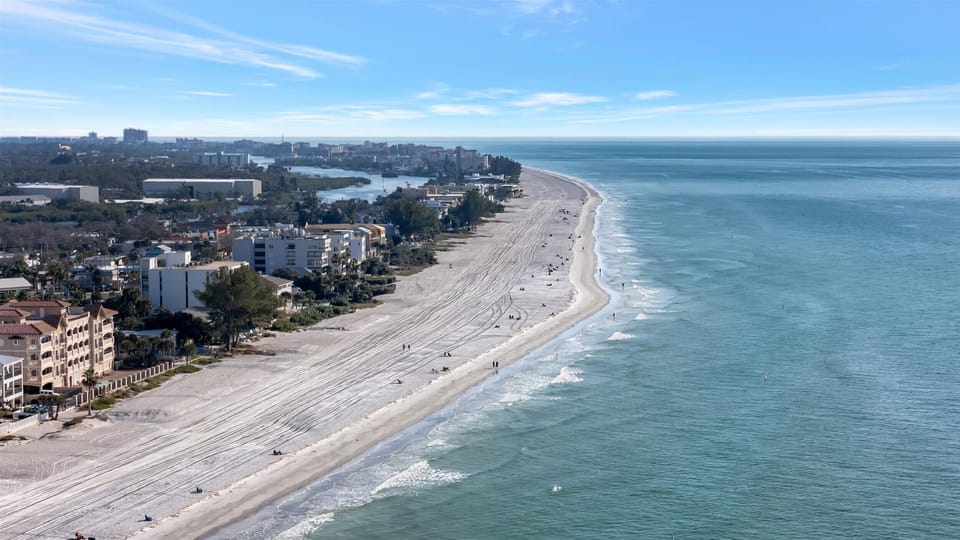 Aerial view showcasing the pristine beachfront stretching along the coastline, with residential buildings nestled among lush greenery near the waterfront area.