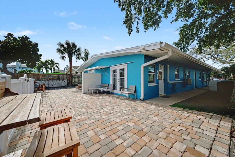 A cheerful turquoise cottage set in a spacious courtyard with brick pavers, palm trees, and shaded seating areas perfect for relaxing outdoors.