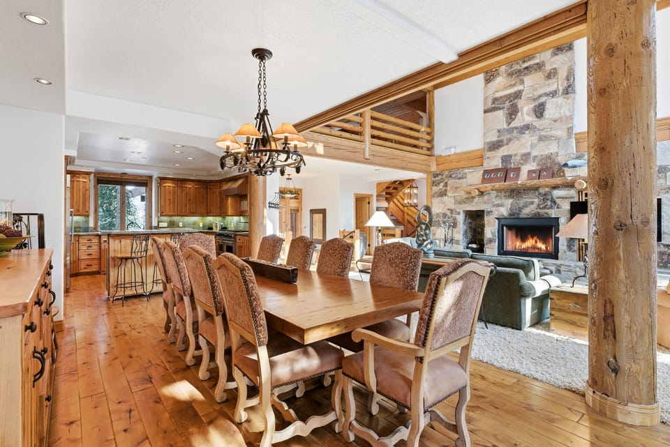 Rustic kitchen and dining space with a large wooden dining table, log cabin accents, chandelier lighting, and stone fireplace in the background.