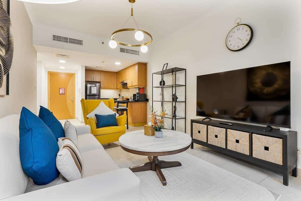 Bright and inviting living room featuring a cozy sitting area, and a 65-inch TV, while the open kitchen seamlessly connects to the living area in the background, creating a spacious and welcoming layout.

