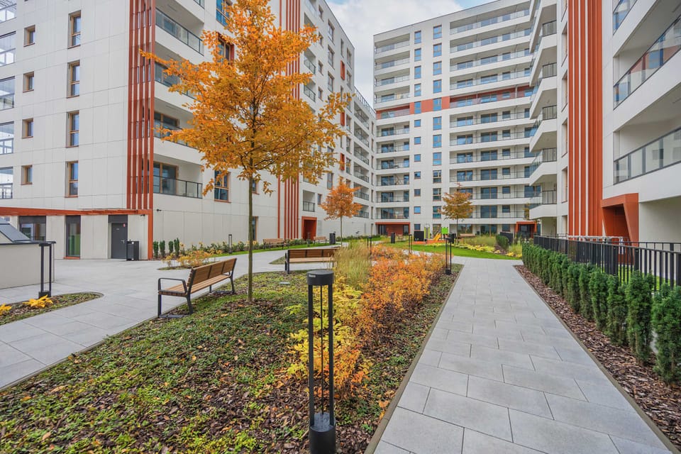 A walkway lined with trees and modern apartment buildings. The surroundings combine urban living with green spaces.