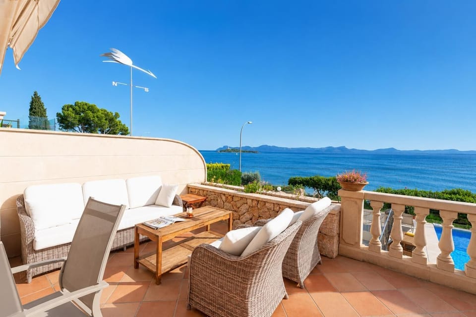 A sunny, tiled terrace featuring comfortable wicker seating, white cushions, and a wooden coffee table overlooking a beautiful blue sea.