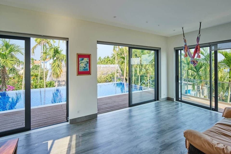 The office space features a large custom wood-slab table, a 27-inch monitor, and a leather sofa, with sunlight from two sliding glass doors doubling as windows, providing views of the river and pool, and room for aerial yoga/ yoga practice.
