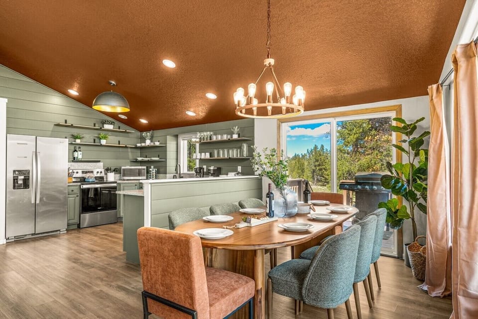 Spacious mountain cabin dining room and kitchen featuring an oval wood table set for six, green and rust upholstered chairs, a ring chandelier, green shiplap accent walls, stainless steel appliances, and large sliding doors opening to a scenic forest deck