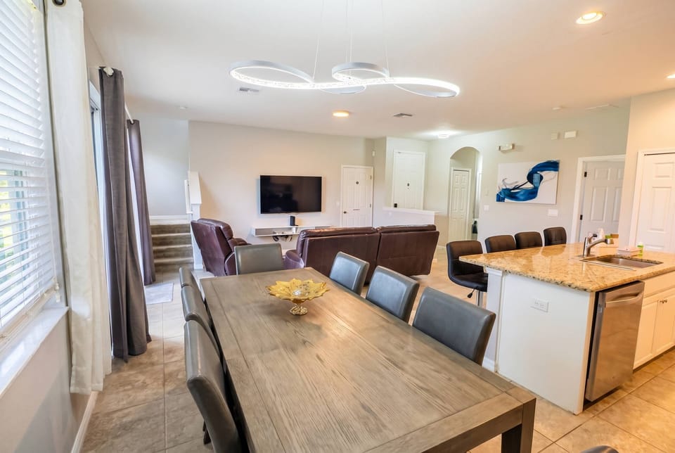 This bright and airy open dining and living area features a sleek wooden table and leather seating, perfect for gatherings. Large windows flood the space with natural light, enhancing its modern decor and inviting ambiance.