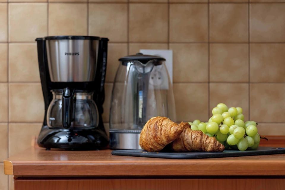 A close-up of a countertop displaying a coffee machine, electric kettle, cups, and a plate of fruit.