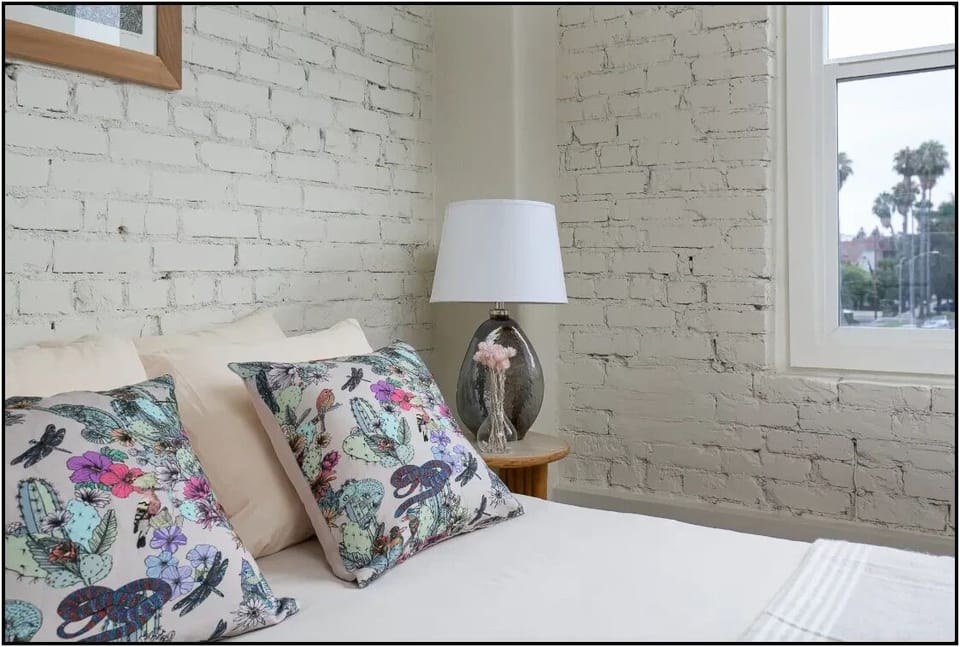 Drift into dreamland in your cozy retreat with charming exposed brick walls and vibrant botanical pillows creating the perfect restful sanctuary.
