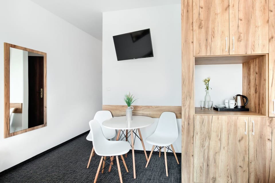 A compact dining-and-sleeping area with a small round table, two white chairs, and a bed positioned beside a tall wooden shelving unit. A wall-mounted TV faces the room.

