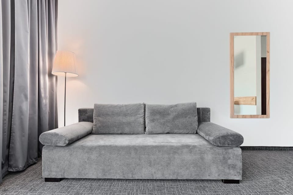 A gray sofa arranged against a clean white wall, paired with a tall mirror and soft lighting. A floor lamp adds a touch of ambience to the simple living corner.

