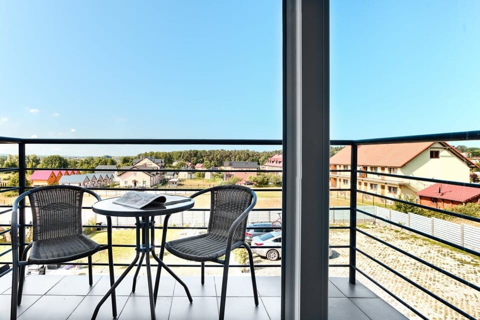An outdoor balcony furnished with a round table and several chairs, overlooking a sunny landscape with rooftops and open sky.