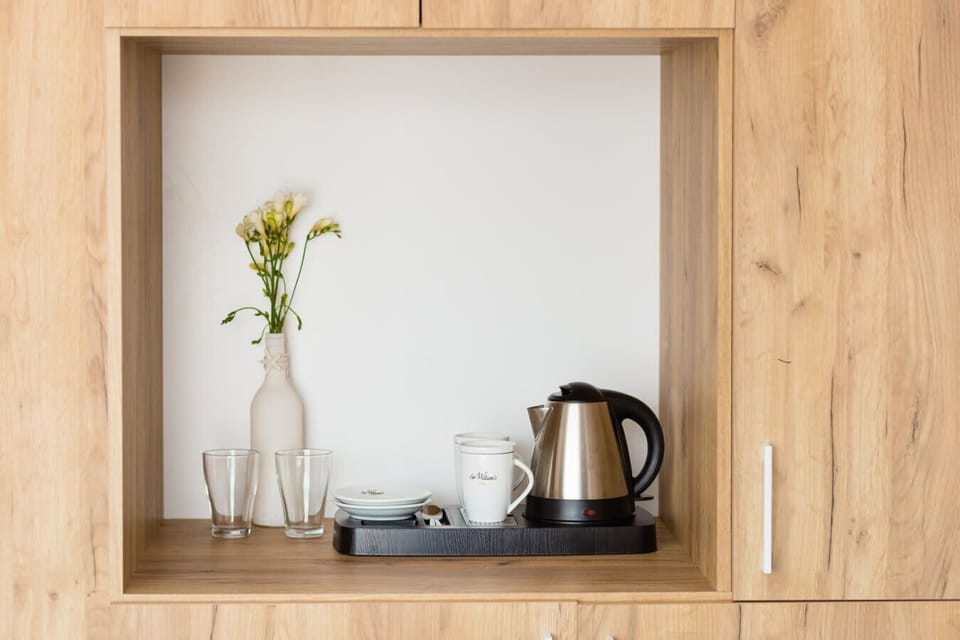 A cozy setup featuring a wooden shelf holding a vase with flowers, a kettle, and cups. The light wood finish adds warmth to the small nook.

