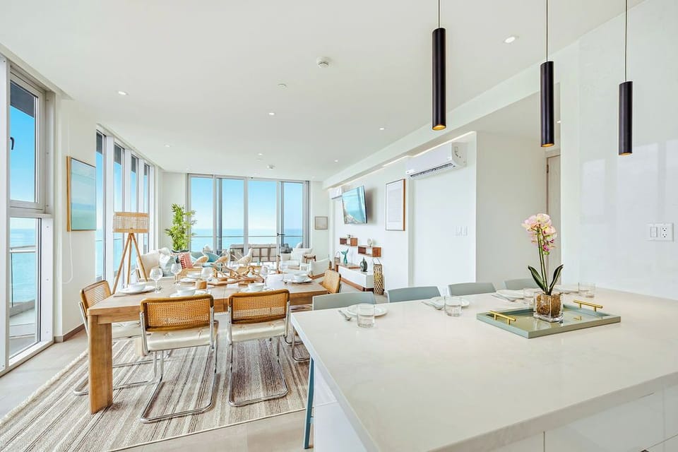 Gather around this elegant dining space with sweeping ocean views through floor-to-ceiling windows. Sunlight fills the room, highlighting modern furnishings and a welcoming table set for your vacation meals.