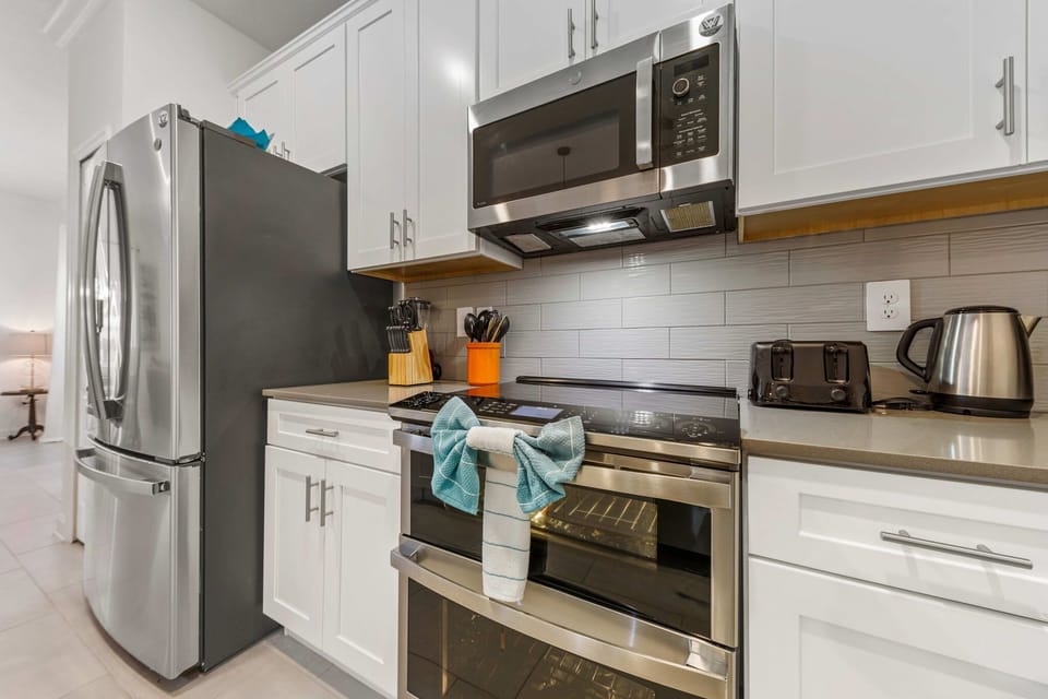 Cook with ease in the modern kitchen-stainless appliances, smooth-top range, microwave, and kettle on sleek quartz counters with subway tile and white shaker cabinets.