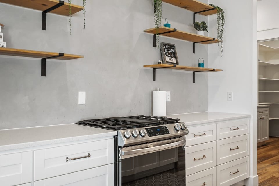 Chef's kitchen with gas range, white cabinetry, and floating shelves.