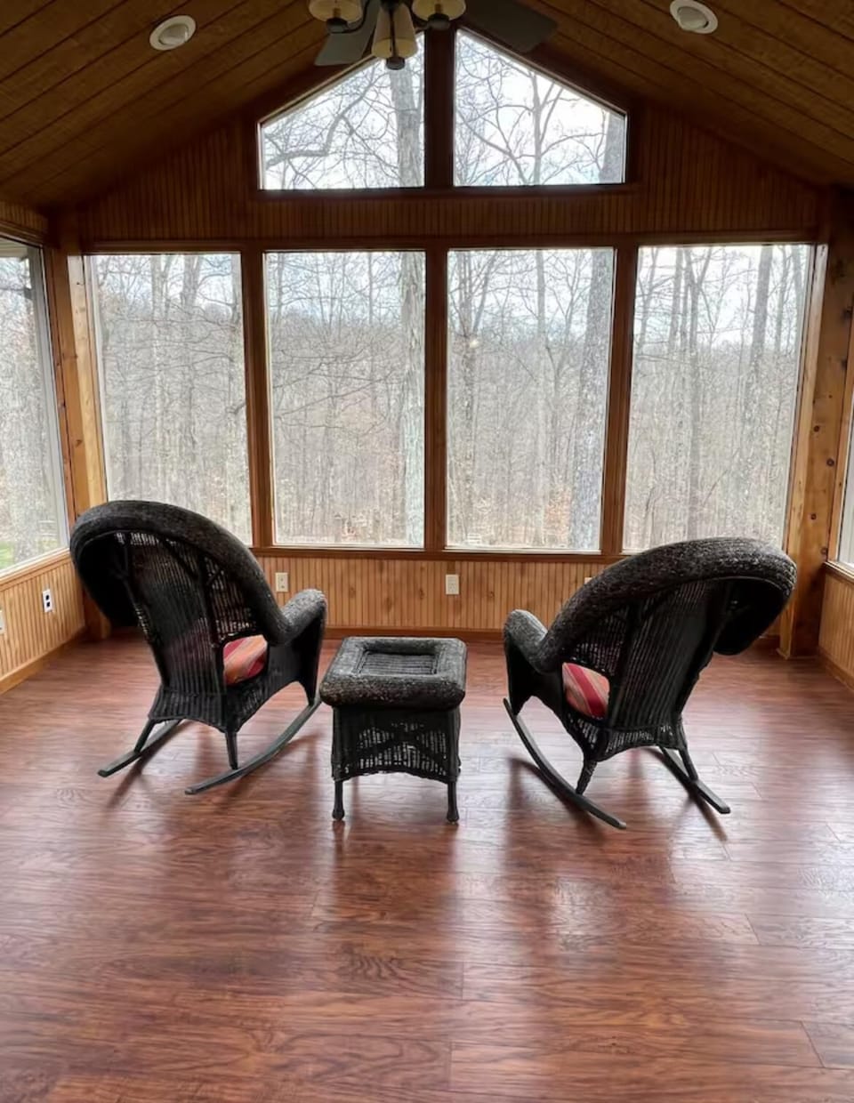 Sunroom with vaulted wood-paneled ceiling, large windows overlooking bare trees, and two black wicker rocking chairs.