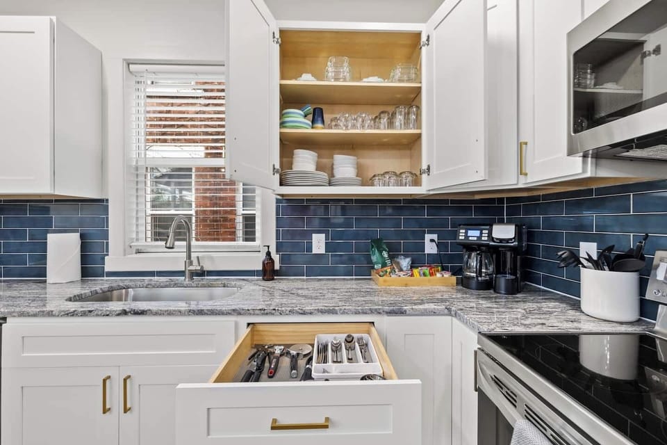 Sleek white cabinetry, a striking blue subway tile backsplash, and granite-style countertops. It comes fully equipped for your cooking needs, including dishes, glassware, a coffee maker, and essential tools, and a full set of utensils.