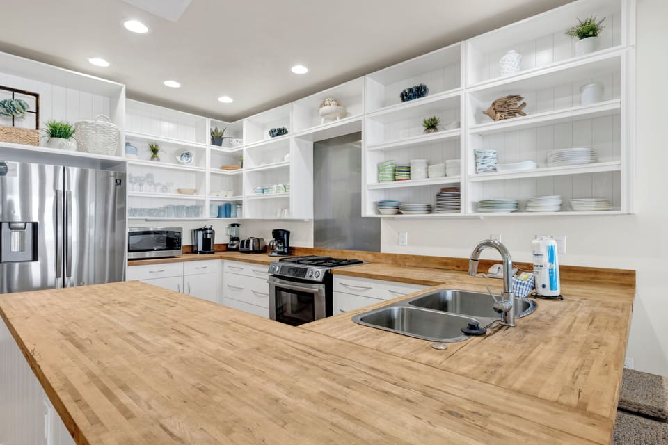 Your gourmet kitchen features modern appliances, butcher-block counters, and open shelving showcasing coastal-themed dishware.