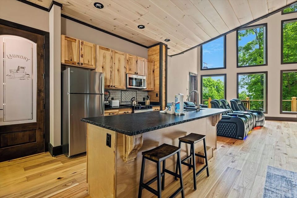 Your open-concept retreat features a fully equipped kitchen with modern appliances and a granite island, flowing seamlessly into a bright living space framed by soaring windows overlooking lush greenery.