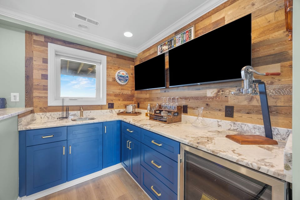 Your private wet bar features bold blue cabinetry, gleaming marble counters, a sink, dual TVs, and mini fridge—perfect for entertaining or unwinding after a beach day.