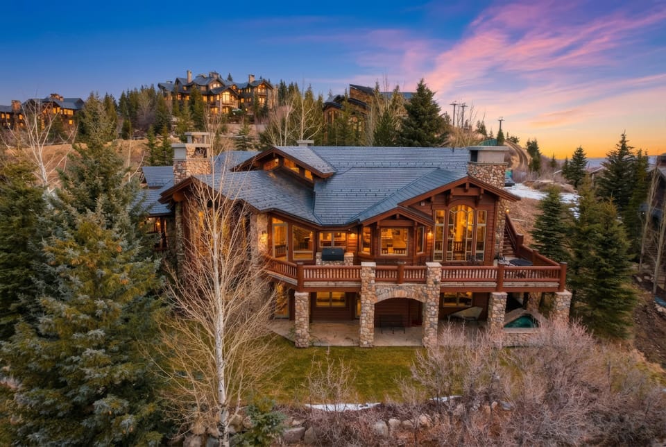 A stately lodge with glowing lights indoors and a sunset sky. you can see the mountains of deer valley in the background and a glowing hot tub