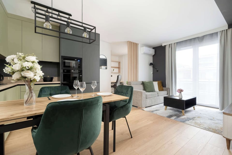 A dining area with a wooden table surrounded by green upholstered chairs. Shelving and decorative lighting add warmth to the space. The room connects seamlessly with the living area.