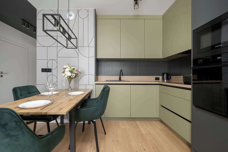 A dining corner with a wooden table and green chairs placed next to the kitchen. Pendant lighting hangs above the table. The setup looks ideal for everyday meals.