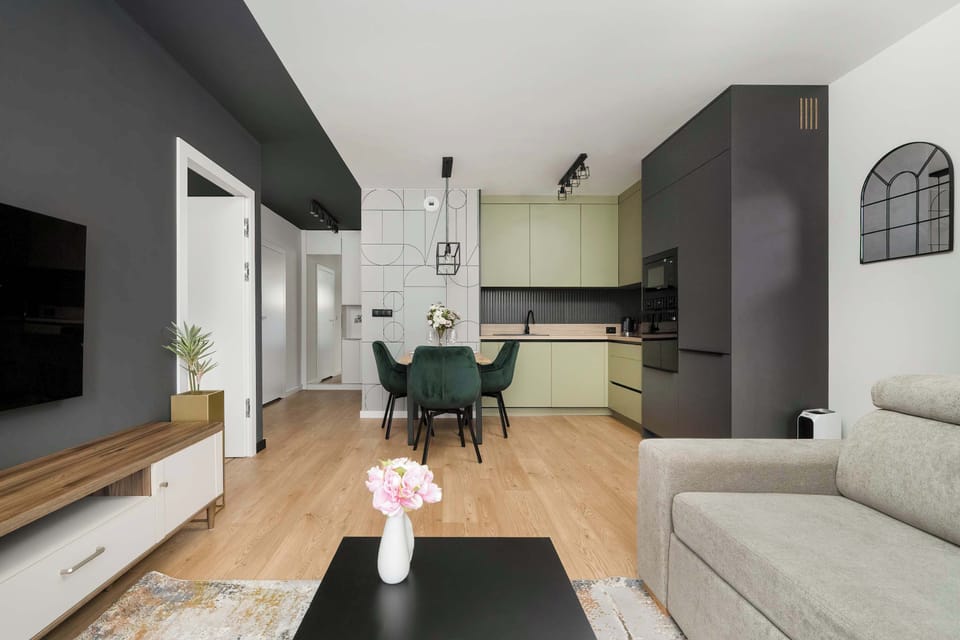 An open-plan living and kitchen area with a sofa, dining table, and kitchen cabinets in the background. A pink flower centerpiece adds a soft decorative touch. The layout feels spacious and functional.