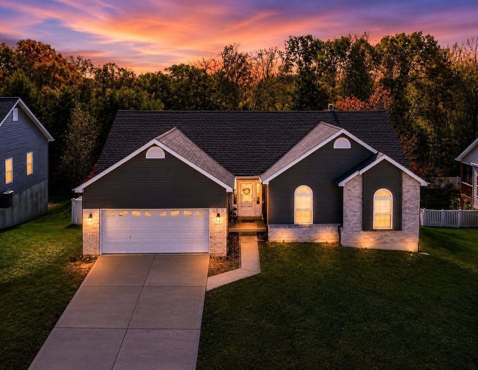 The illuminated single-story home and two-car garage stand against a colorful sunset and wooded backdrop.