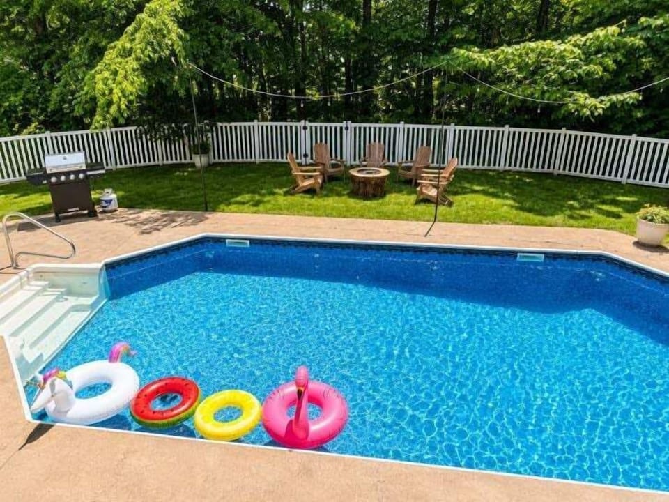 The clear blue pool is framed by a spacious deck, colorful floats, Adirondack chairs, and a nearby firepit.