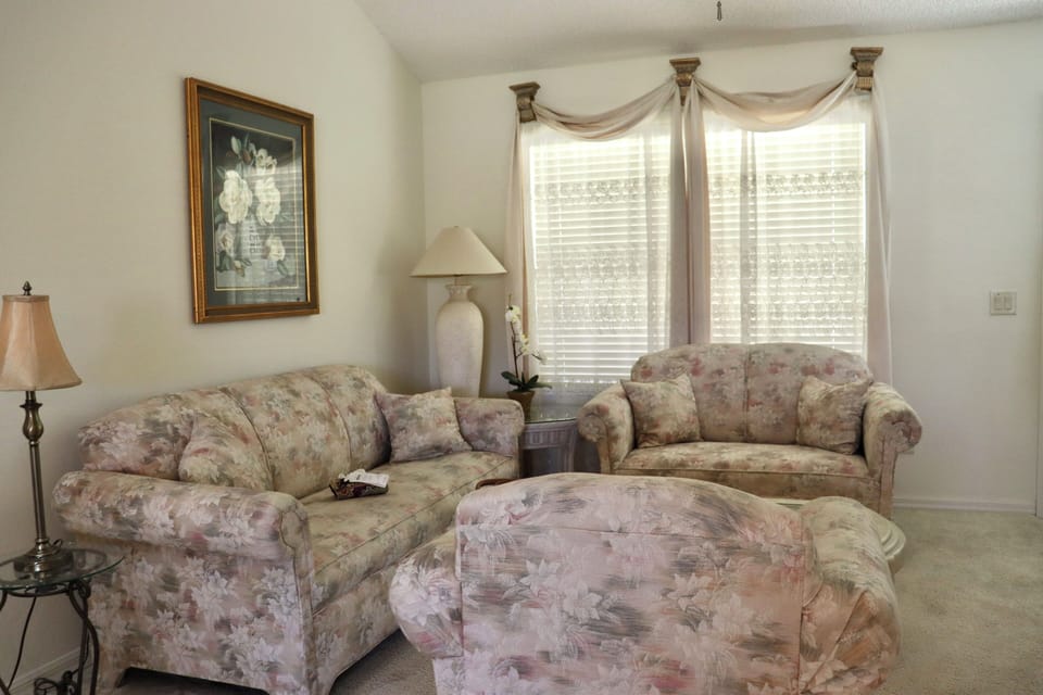 Relax in your elegant living room with plush floral seating, soft lighting, and cheerful windows that fill the space with natural light.