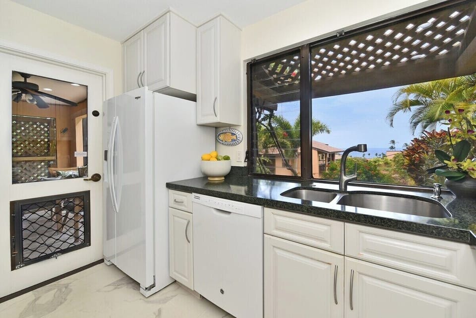 A bright kitchen with white cabinetry, dark granite countertops, and wide windows framing lush garden views.