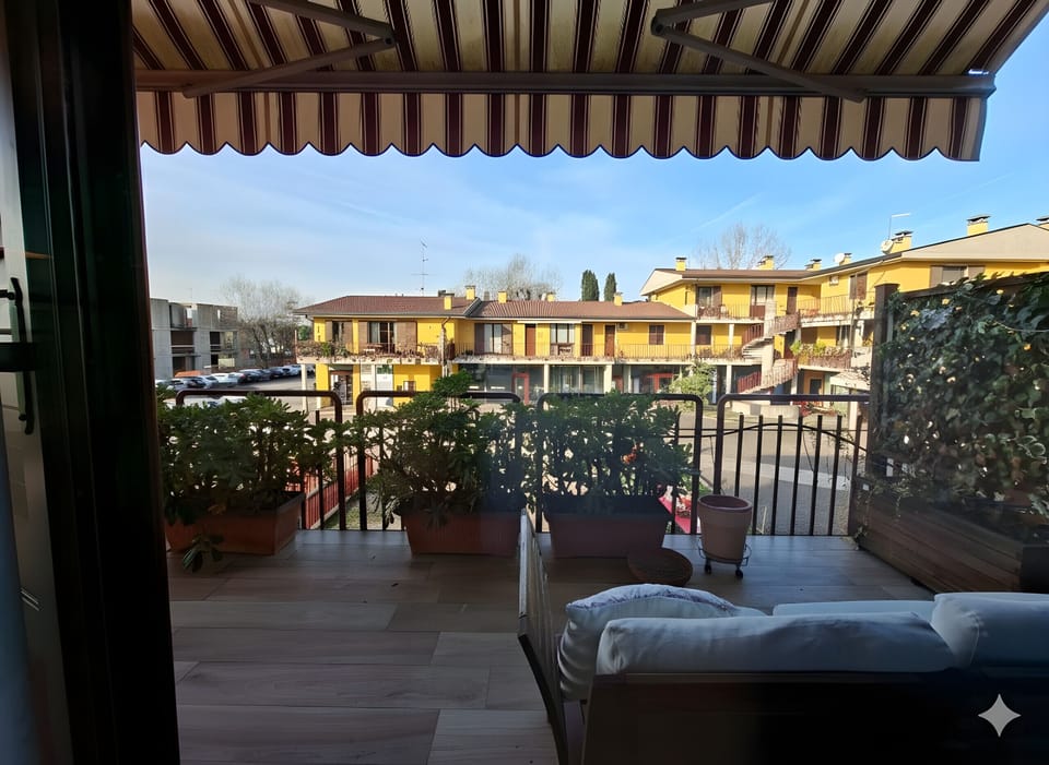 Covered balcony of the apartment located in Peschiera del Garda, equipped with an awning, seating, and decorative plants. The outdoor space offers a pleasant view of the surrounding residential area and is ideal for relaxing outdoors at any time of day.