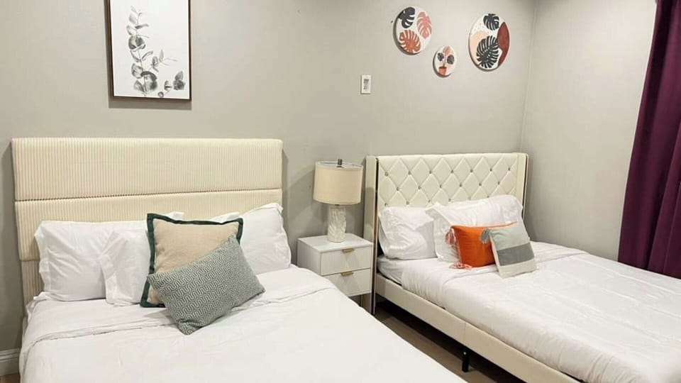 A spacious, stylish bedroom featuring two cozy beds with plush upholstered headboards, soft layered pillows, and modern decor. Warm lighting, elegant wall art, and rich curtains create a comfortable, inviting space perfect for families or groups.
