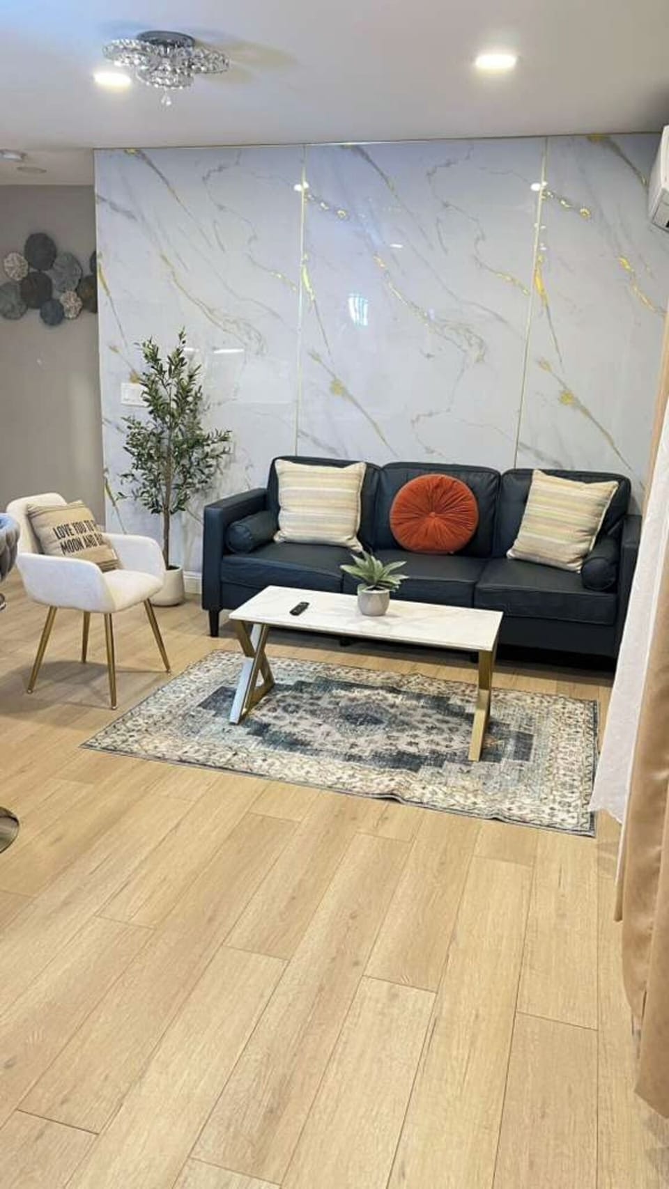 A cozy, modern living room featuring a sleek dark sofa, chic accent chair, marble‑panel wall, and warm lighting. Stylish decor, a contemporary rug, and a clean coffee table create an inviting space perfect for relaxing or entertaining guests.