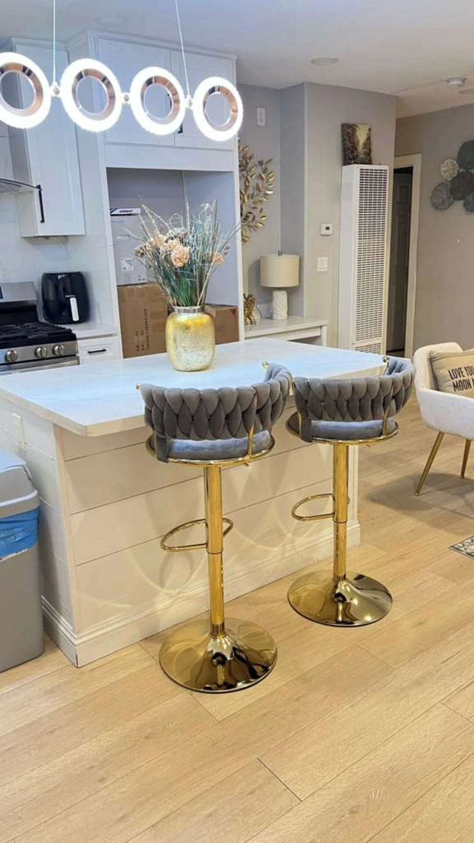 A bright modern kitchen with a spacious island, elegant grey velvet bar stools, stylish ring lighting, and sleek white cabinetry. Stainless‑steel appliances and warm wood floors create a clean, inviting space ideal for cooking and gathering.