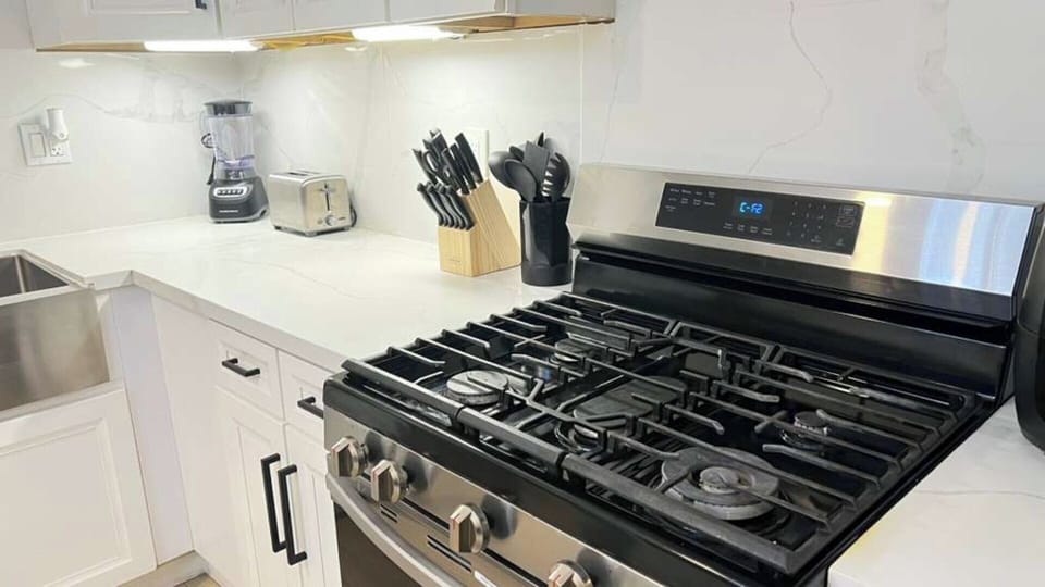 A bright, modern kitchen with sleek white countertops, stainless‑steel gas stove, blender, toaster, and a full knife set. Clean finishes, under‑cabinet lighting, and organized essentials make this space perfect for cooking and everyday convenience.