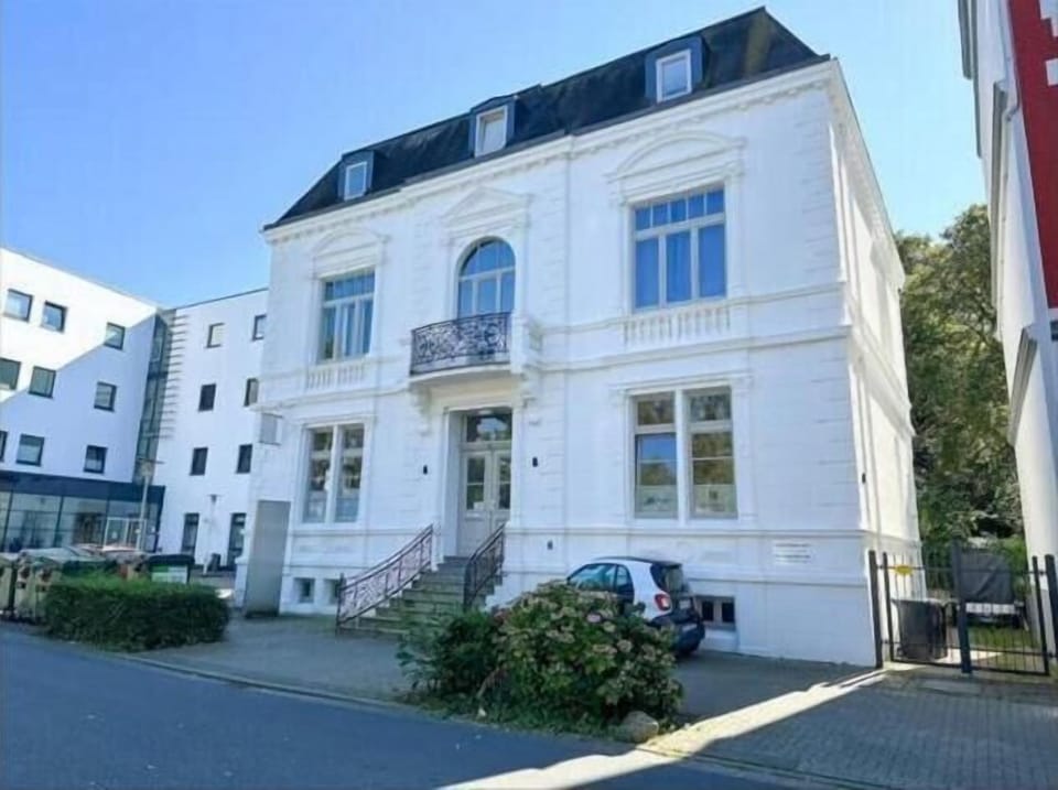 Diese prachtvolle weiße Villa in Wilhelmshaven besticht durch ihre historische Architektur. Filigrane Balkongitter und hohe Fenster verleihen dem Gebäude an der Nordseeküste einen edlen Charme, der unter blauem Himmel besonders strahlt.