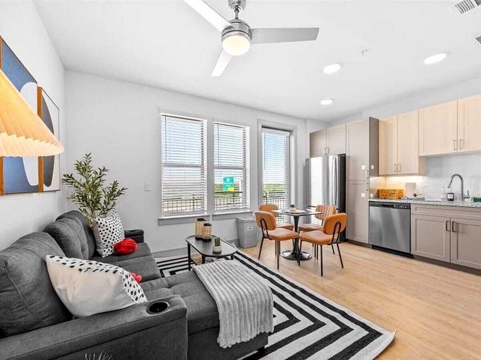 Bright and stylish living room featuring a modern gray sectional, bold black-and-white rug, and warm wooden dining set beside large windows that fill the space with natural light.
