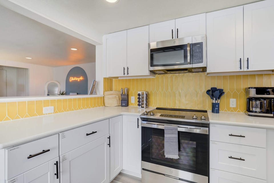 The fully stocked kitchen features stainless steel appliances, a modern gold tile backsplash, and sleek white cabinetry. It is thoughtfully equipped with everything needed to prepare home-cooked meals during your stay.