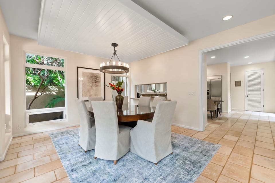 Your elegant dining space features a stunning round table for eight beneath a dramatic chandelier, with tropical garden views through expansive windows.