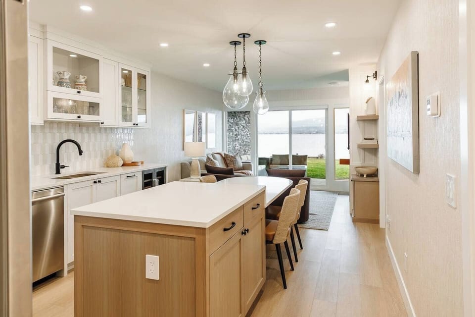 Modern kitchen with white cabinets, a white countertop, black bar stools, and tiled flooring, with a dining area in the background.