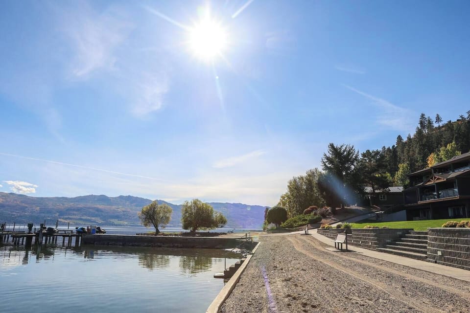 A serene lakeside scene with a stone fire pit filled with white rocks, set against calm waters and rolling hills, under a clear blue sky.


