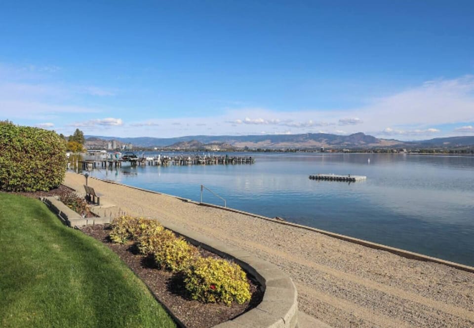 Lakeside pathway lined with shrubs and flowers, leading to a dock with boats and a mountain range across the water.
