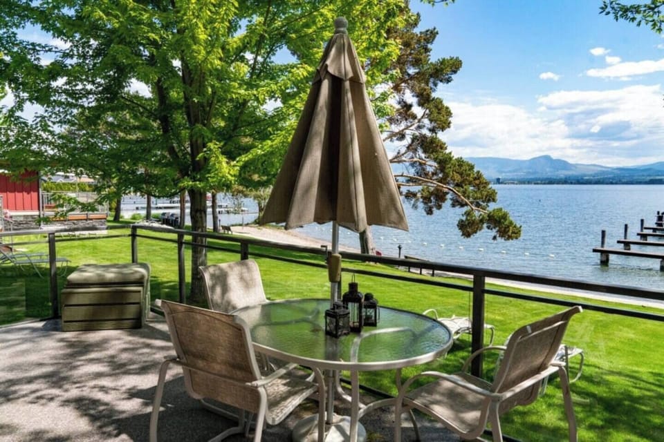 Balcony with a table, chairs, and a green umbrella, overlooking a serene lake surrounded by lush greenery.
