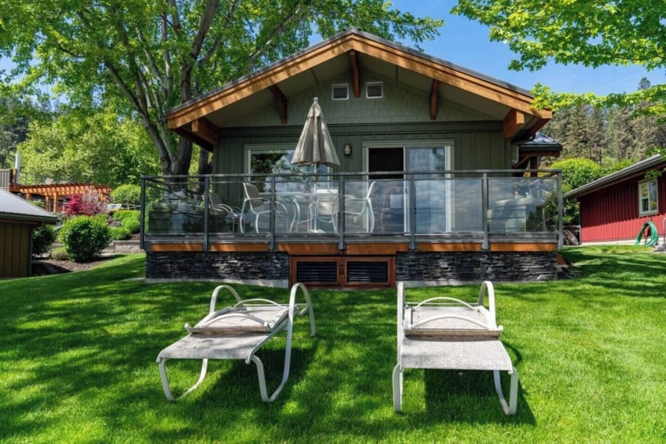 Cozy cabin with a wooden facade, glass-railed balcony, and lounge chairs overlooking the lake.




