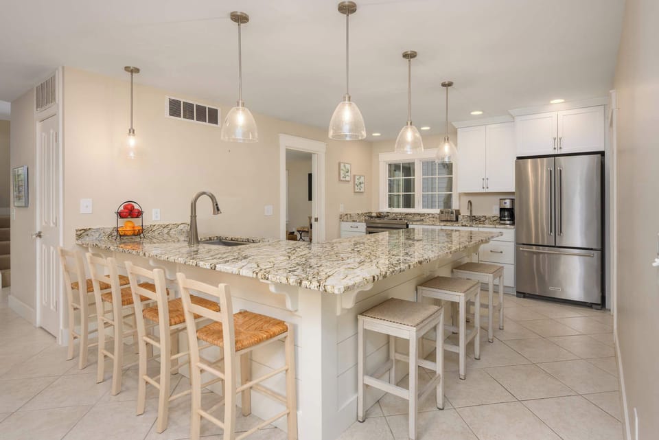 A gourmet kitchen highlighted by a massive granite-topped peninsula with seating for six. Elegant pendant lighting hangs above the breakfast bar, while the stainless steel refrigerator and bright finishes create a sophisticated, clean aesthetic.
