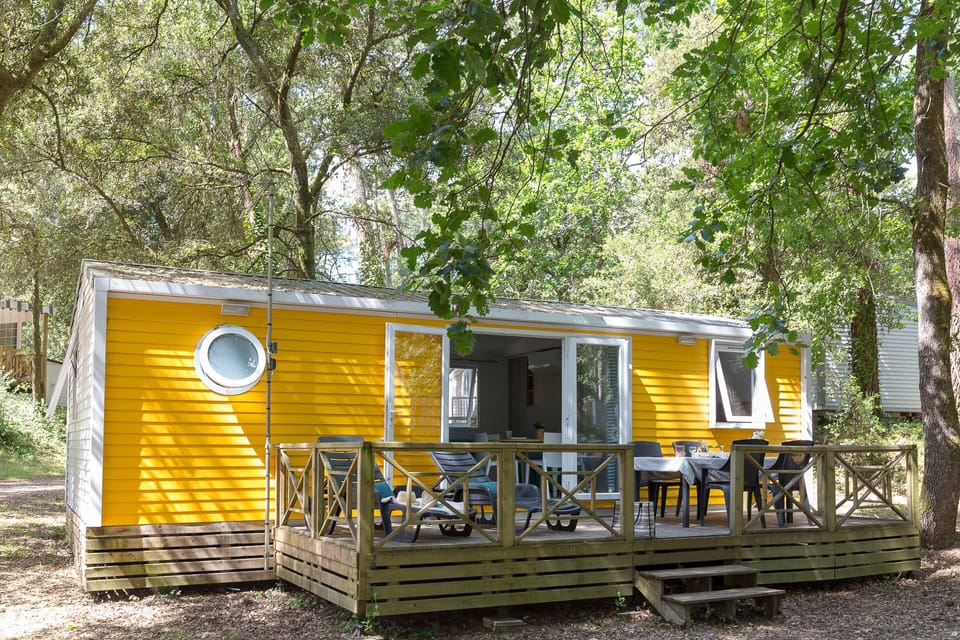 The vibrant yellow mobile home boasts a charming wooden terrace, perfect for enjoying meals outdoors in the serene surroundings of Camping le Logis.