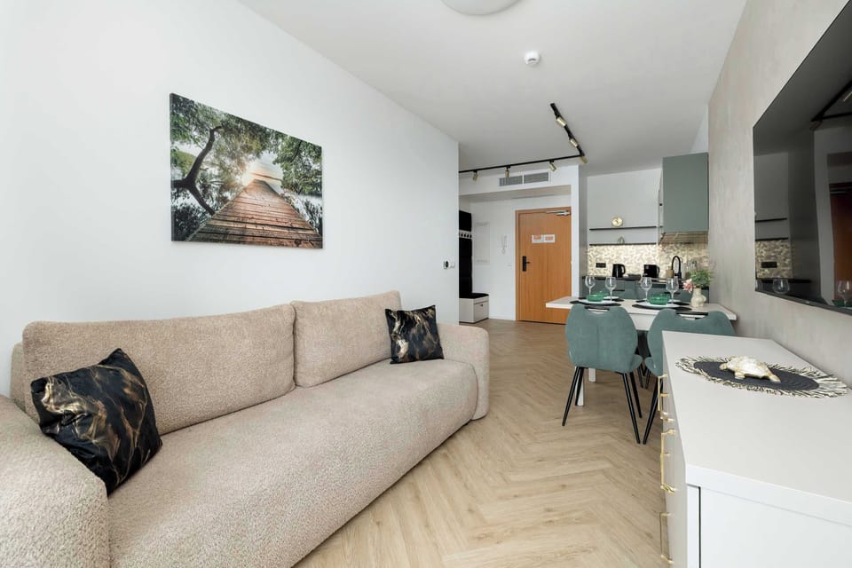 A bright living area with a beige sofa and a dining table placed near the kitchen. Soft lighting and neutral tones create a welcoming atmosphere. The open layout connects all zones smoothly.