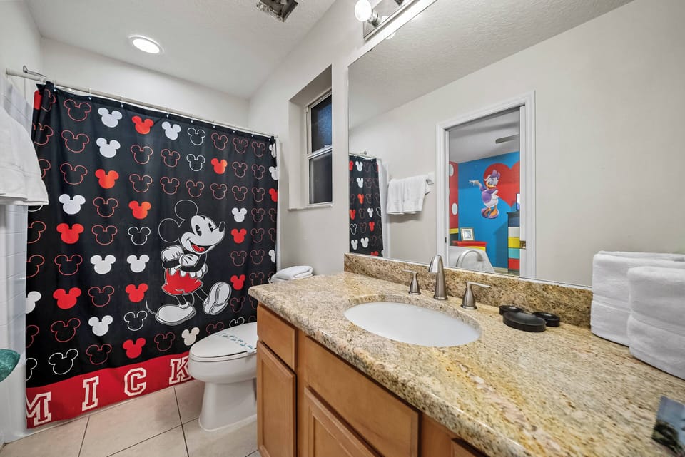 Step into a playful bathroom featuring a vibrant Mickey Mouse shower curtain that adds a touch of whimsy. The warm wood cabinetry complements the sleek granite countertop, while natural light brightens the inviting space.